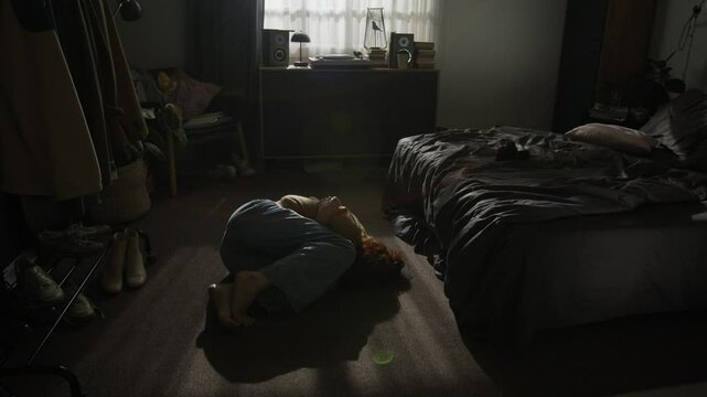 Unrecognizable distressed woman lying curled on floor of sunlit bedroom, near bed and scattered belongings