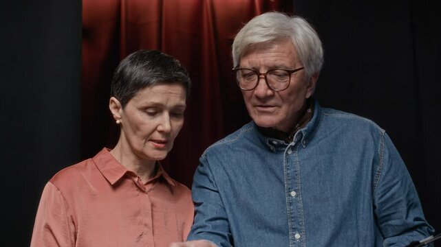 Tilt up shot of senior Caucasian director discussing script with mature female screenwriter standing against red theatre curtains