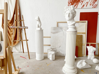 A bright art studio with white sculptures on columns. The background features easels, canvases, and draped fabric in warm colors. A creative atmosphere is present.