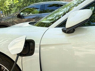 A close-up view of a white electric car charging. The charging port is open, and a charging cable is connected. A dark vehicle is visible in the background.