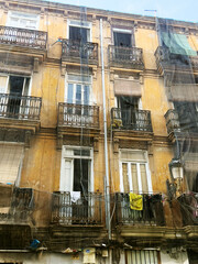 An old yellow apartment building with multiple balconies. Clothes hang on the railings. Windows are partially open, revealing a glimpse of the interior.