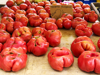 A variety of ripe, red heirloom tomatoes displayed on a wooden table at a market. Some tomatoes have unique shapes and sizes, showcasing their organic nature.