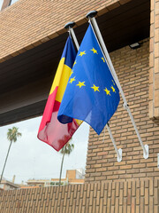Two flags displayed on a building. The left flag is the Romanian flag with yellow, blue, and red stripes. The right flag is the European Union flag with a blue background and yellow stars.