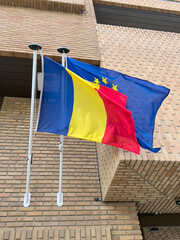 Flags of Romania and the European Union waving in the wind against a brick building background. Bright colors and clear design are visible.