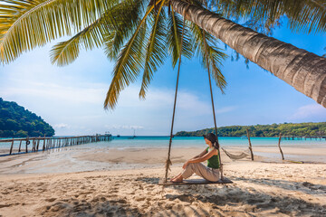 Happy traveler asian woman with cap relax in swing on sea beach at Koh Kood Trat Thailand Summer travel holiday vacation concept