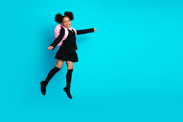 Joyful schoolgirl jumping excitedly while wearing a backpack and formal uniform against a solid teal background