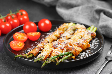 Grilled asparagus wrapped in crispy bacon sprinkled with parmesan cheese served on plate with fresh cherry tomatoes on black background. Gourmet dish, cooking recipe, cuisine, or restaurant menu
