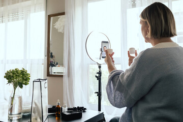 Middle-aged woman beauty blogger filming skincare product unboxing at home with ring light....