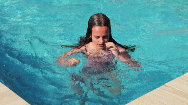A young girl with wet hair and closed eyes emerges from the clear blue water of a swimming pool, enjoying the sun.