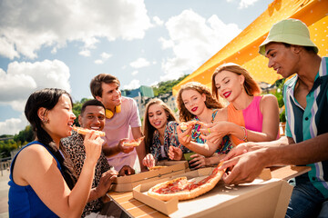 Group of cheerful young friends enjoying pizza outdoors on a sunny day while sharing smiles and...