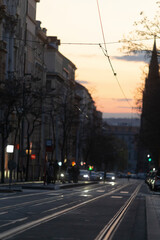 A bustling urban street scene at dusk, showcasing a variety of buildings, extensive tram tracks, and numerous vehicle lights glimmering under a beautifully painted twilight sky