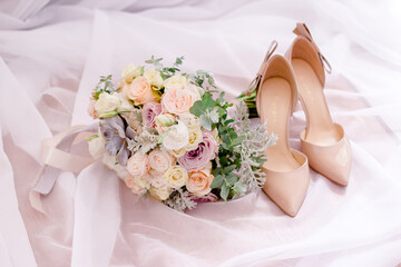 A beautiful bridal bouquet of pastel roses, eustoma, and a succulent lies next to a pair of elegant...