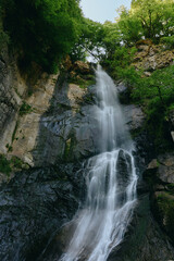 Obraz premium Stunning waterfall cascades down rocky cliffs surrounded by lush green forest and vibrant foliage, creating a refreshing and serene natural scene in daylight.