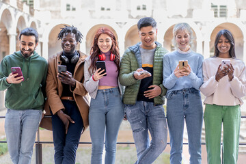 Diverse group of young adults standing in a row using smartphones in urban setting with joyful...