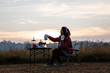 Fototapeta premium woman sitting outdoors with camping gear, including a table, chair, and a steam lamp. She is enjoying her morning coffee in a serene, natural setting.
