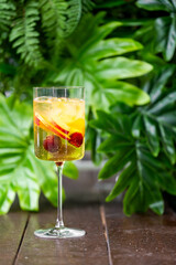 A tall glass of refreshing cocktail with ice cubes, apple slices, and cherries, set against a lush green tropical background, creating a vibrant and inviting summer atmosphere.