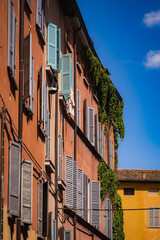 Reggio Emilia, Italy, 01.04.2025. Reggio Emilia, Italy, 01.04.2025. Rusty red facade with pastel shutters, climbing green ivy and clear blue sky in old town street