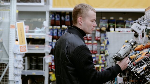 Construction worker comparing cordless drill features and prices while shopping in a hardware store, examining design and specifications. A man in a hardware store choosing a tool.
