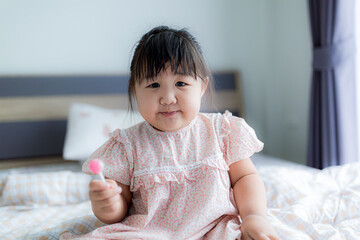 Fototapeta premium A cute little girl is happily eating a lollipop,Sweet tooth Asian girl happy holding big lollipop sitting on bed at home