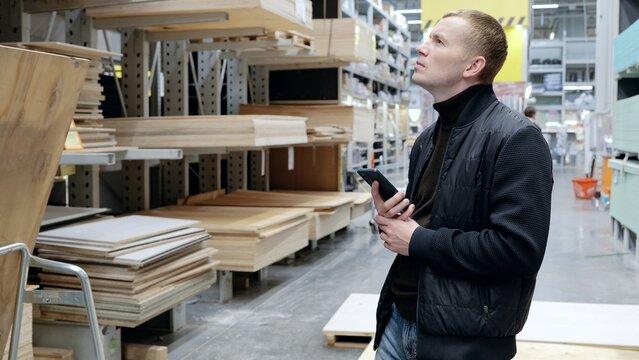 Contractor using a smartphone to select wood materials in a lumber warehouse for an upcoming project. A man in a hardware store, choosing lumber, shelves with lumber.
