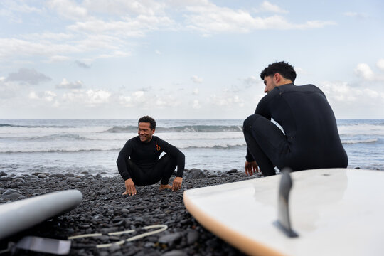 Surfers wearing wetsuits crouching on pebble beach and looking at ocean waves - Powered by Adobe