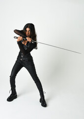 Obraz premium Fantasy female warrior in leather armour full body studio portrait of fierce brunette in catsuit with daggers and sword, posing as assassin, rogue, knight or storybook character on white background