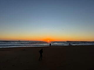 Sunset over the ocean at the beach with silhouettes of people walking along the shore during...