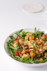 A fresh, colorful salad featuring arugula, avocado, cherry tomatoes, quinoa, roasted squash, and almond slices in a white bowl.