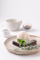 A decadent chocolate brownie topped with vanilla ice cream and garnished with mint and powdered sugar on a rustic plate, with blurred coffee cup and creamer in the background.