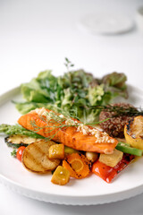 Grilled salmon fillet garnished with herbs, served with roasted vegetables and leafy greens on a white plate.