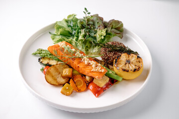 A healthy, colorful plate featuring grilled salmon, mixed roasted vegetables, fresh leafy greens, and a charred lemon half on a white dish.