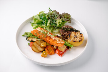 A plated dish featuring grilled salmon, mixed greens, roasted vegetables, quinoa, and a charred lemon half on a white plate.