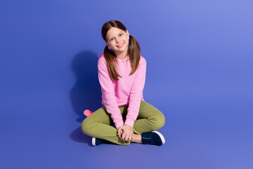 Charming schoolgirl sitting cross-legged on purple background wearing a pink hoodie and green pants...