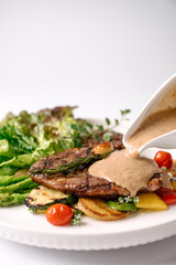 Grilled steak with creamy sauce being poured, served with asparagus, cherry tomatoes, zucchini, and greens on a white plate.