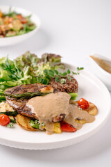 A gourmet steak dish served with creamy sauce, grilled vegetables, and quinoa, accompanied by a fresh green salad in the background.