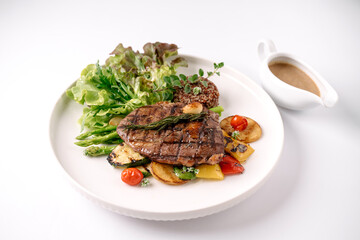 Grilled steak served with fresh leafy greens, roasted vegetables, and a side of creamy sauce in a white dish on a white background.