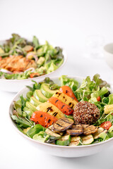 Two fresh, colorful salad bowls featuring grilled vegetables, avocado slices, quinoa, and mixed greens on a white background.