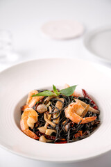 Black squid ink pasta with shrimp, mushrooms, and herbs served in a white bowl on a white table.