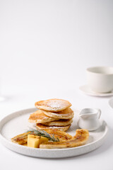 A stack of fluffy pancakes dusted with powdered sugar, served with caramelized banana slices, butter cubes, and a small pitcher of syrup on a white plate.