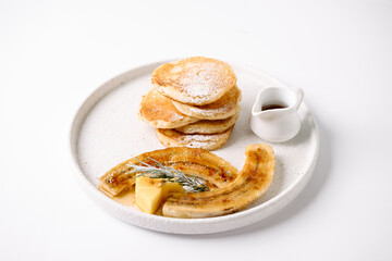 A stack of golden pancakes dusted with powdered sugar, served with caramelized banana halves, a small block of butter, rosemary, and a side of syrup on a white plate.