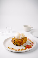 A warm dessert bread topped with two scoops of vanilla ice cream, garnished with a strawberry and crumbs on a white plate with a minimalist background.