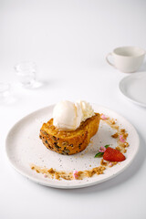 A plated dessert featuring a slice of bread pudding topped with a scoop of vanilla ice cream and whipped cream, garnished with strawberry and crumbs.