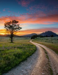 Scenic Dirt Road Leading Through a Grassy Meadow Towards Distant Mountains Under a Vibrant Sunset Sky - Landscape Photography