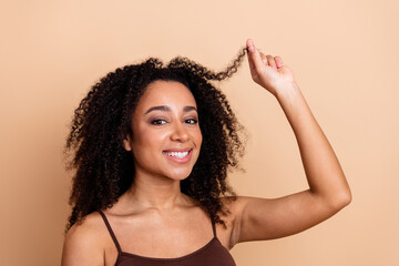 Young confident woman with curly hair smiling and touching her locks against a beige background...