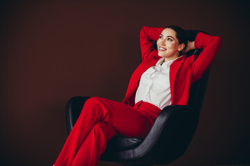 Gorgeous Confident Businesswoman in Red Suit Relaxing on Chair Against Brown Background