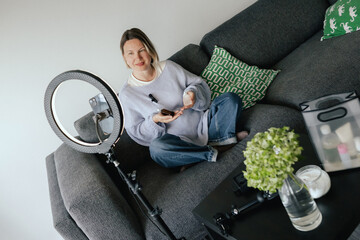 Middle-aged woman beauty blogger filming skincare product unboxing at home with ring light....