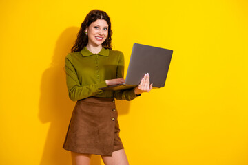 Fototapeta premium Cheerful young woman holding a laptop against a vibrant yellow background while showcasing modern casual style