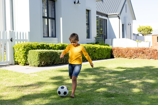 Barefoot child kicking black-and-white soccer ball across green lawn near white house and hedges