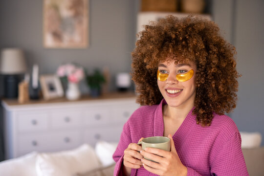 Cheerful Woman in Pink Robe with Eye Patches Relaxing and Holding Mug in Cozy Room - Powered by Adobe