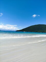 Obraz premium A scenic view of a Ubatuba tropical beach with white sand, blue water, and a lush green hill under a clear blue sky
